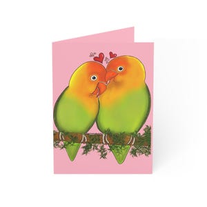 May include: A folded greeting card with a cartoon illustration of two lovebirds. The birds have orange and yellow heads, green bodies, and red beaks. The card is set against a pink background, with small red hearts floating above the birds.