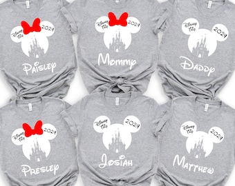 May include: Five grey t-shirts featuring a white silhouette of Cinderella's castle and Mickey Mouse ears. Each shirt has a red bow and the text "Disney Trip 2024". The shirts are personalised with the names "Paisley", "Mommy", "Daddy", "Presley", and "Josiah".