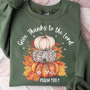 May include: Green sweatshirt with a stacked pumpkin design. The design includes a cream-colored pumpkin, a leopard print pumpkin, and an orange pumpkin with floral accents. The text "Give Thanks to the Lord" and "Psalm 136:1" are also featured.