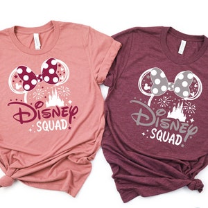 Puede incluir: Dos camisetas con una silueta blanca de orejas de Mickey Mouse con un lazo y el texto "Disney Squad" en blanco con un castillo rosa y fuegos artificiales. La camiseta de la izquierda es de color rosa polvoriento y la camiseta de la derecha es de color burdeos.