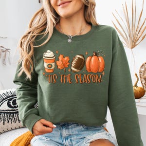 Puede incluir: Sudadera verde con un gr&aacute;fico de una taza de caf&eacute;, una hoja, una pelota de f&uacute;tbol y una calabaza. El texto "'Tis the season" est&aacute; debajo del gr&aacute;fico.