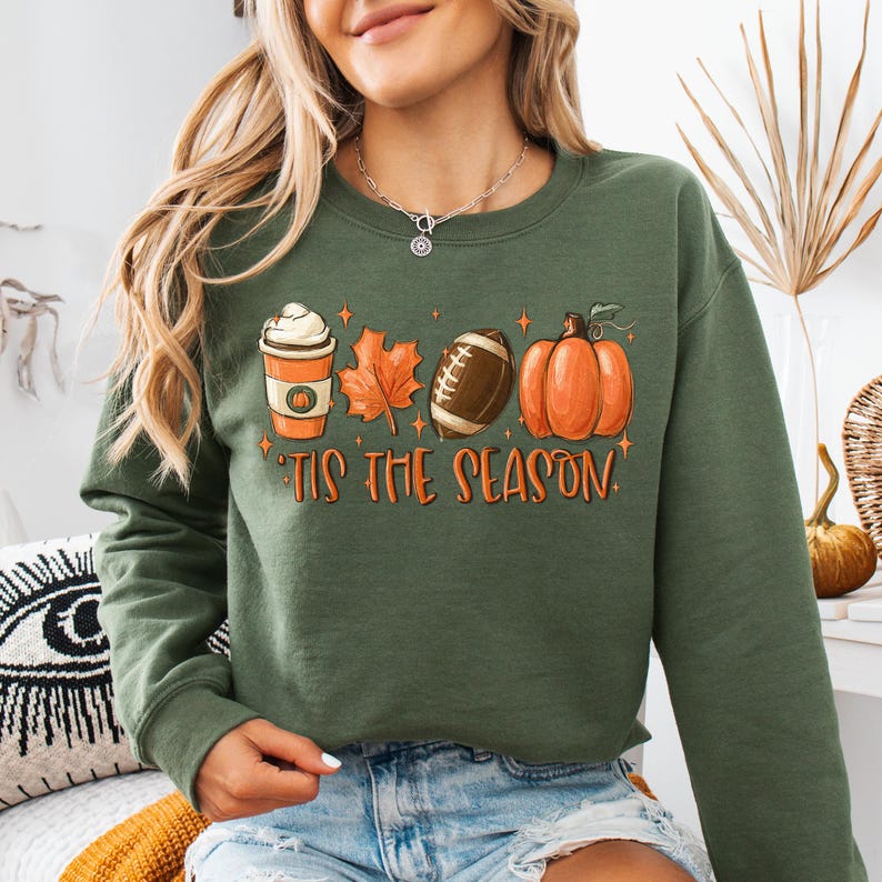 Puede incluir: Sudadera verde con un gr&aacute;fico de una taza de caf&eacute;, una hoja, una pelota de f&uacute;tbol y una calabaza. El texto "'Tis the season" est&aacute; debajo del gr&aacute;fico.