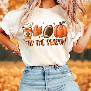 Puede incluir: Camiseta blanca con un dise&ntilde;o gr&aacute;fico de una taza de caf&eacute;, un bal&oacute;n de f&uacute;tbol, una calabaza y hojas de oto&ntilde;o. El texto "Tis the Season" est&aacute; impreso debajo del gr&aacute;fico.