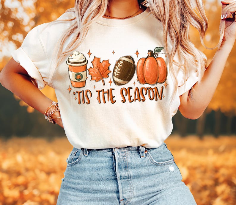 Puede incluir: Camiseta blanca con un dise&ntilde;o gr&aacute;fico de una taza de caf&eacute;, un bal&oacute;n de f&uacute;tbol, una calabaza y hojas de oto&ntilde;o. El texto "Tis the Season" est&aacute; impreso debajo del gr&aacute;fico.