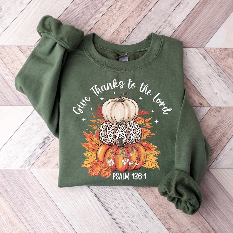 May include: Green sweatshirt with a stacked pumpkin design. The design includes a cream-colored pumpkin, a leopard print pumpkin, and an orange pumpkin with floral accents. The text "Give Thanks to the Lord" and "Psalm 136:1" are also featured.
