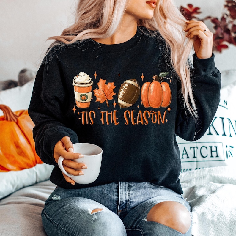 Puede incluir: Sudadera negra con un gr&aacute;fico de una taza de caf&eacute;, una hoja, una pelota de f&uacute;tbol y una calabaza. El texto "Tis the season" est&aacute; impreso debajo del gr&aacute;fico.