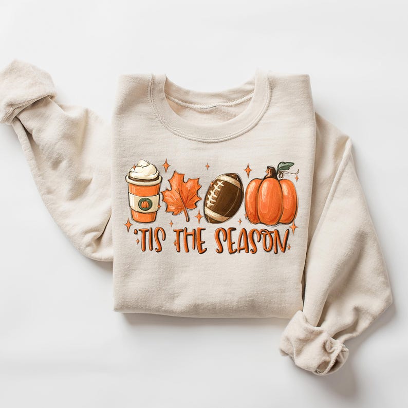 Puede incluir: Una sudadera beige con un dise&ntilde;o gr&aacute;fico de una taza de caf&eacute;, una pelota de f&uacute;tbol, una hoja y una calabaza. El texto "'Tis the season" est&aacute; impreso debajo del gr&aacute;fico.