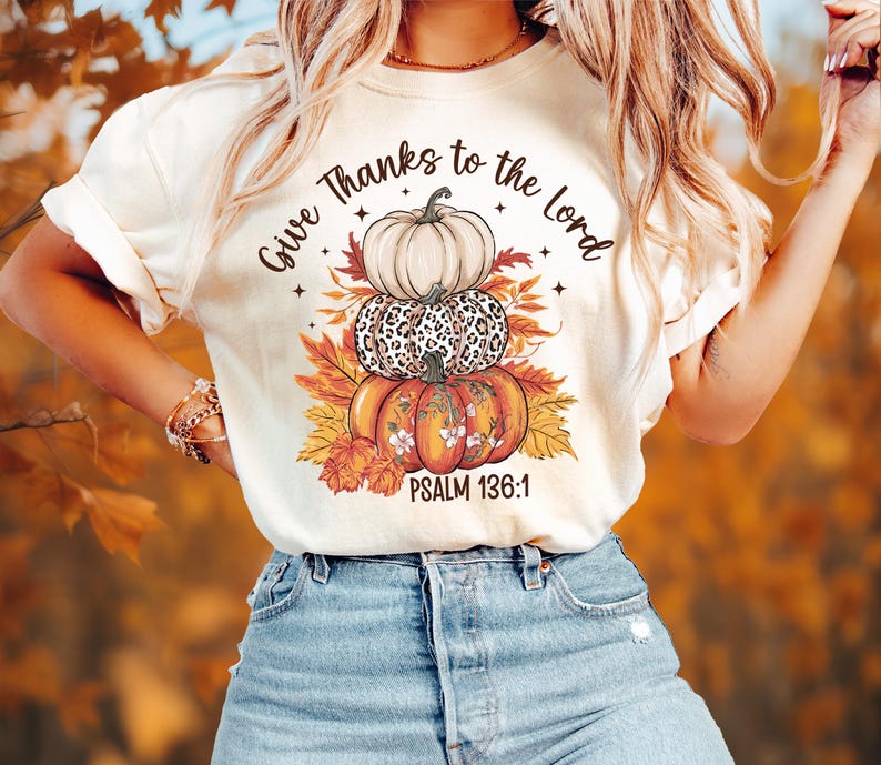 May include: Cream-colored t-shirt featuring a stacked pumpkin design with the text "Give Thanks to the Lord" and "Psalm 136:1". The pumpkins are white, leopard print, and orange, with fall leaves.