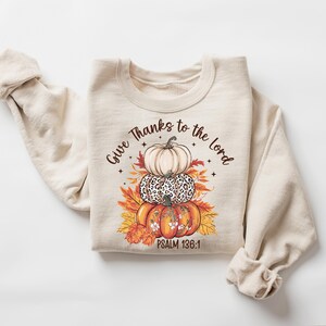 May include: Beige sweatshirt featuring a stacked pumpkin design with the text "Give Thanks to the Lord" and "Psalm 136:1". The pumpkins are white, leopard print, and orange, with fall leaves.