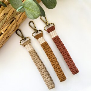 May include: Three macrame wristlet keychains in shades of tan, brown, and rust. Each keychain features a woven cord wrist strap and a bronze-colored clasp. The keychains are arranged diagonally on a white surface.