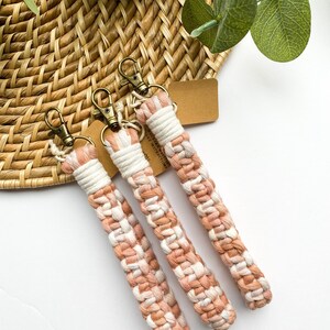 May include: Three macrame keychains in shades of peach, white, and tan. Each keychain features a woven design with a bronze-colored clasp and a small tag. The keychains are arranged on a woven basket.