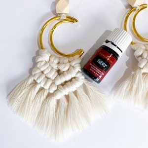 May include: Cream-colored macrame car air fresheners with gold-colored crescent-shaped metal accents and wooden beads. A small bottle of essential oil with the text "THIEVES" is also in the image.