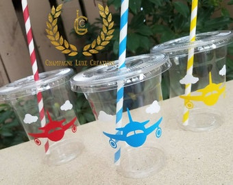 May include: Three clear plastic cups with lids and straws. Each cup features a different colored airplane graphic: red, blue, and yellow. The straws are striped in red and white, blue and white, and yellow and white. The cups are decorated with white cloud graphics.