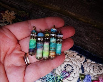 May include: Five handmade beaded pendants with colorful wrapped thread bodies. Each pendant has a different colored bead on top, with decorative metal caps and accents. The pendants are held in a hand.