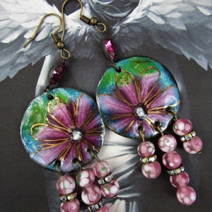 May include: A pair of dangle earrings with pink and purple floral designs on a blue and green background. The earrings have pink and white glass beads with rhinestones hanging from them.