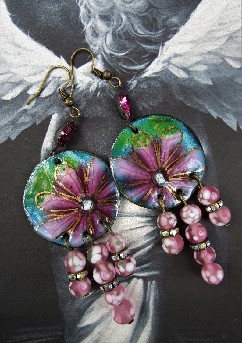May include: A pair of dangle earrings with pink and purple floral designs on a blue and green background. The earrings have pink and white glass beads with rhinestones hanging from them.