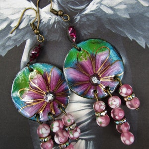 May include: A pair of dangle earrings with pink and green floral designs on the round pendant. The pendants are attached to a chain of pink and white glass beads.