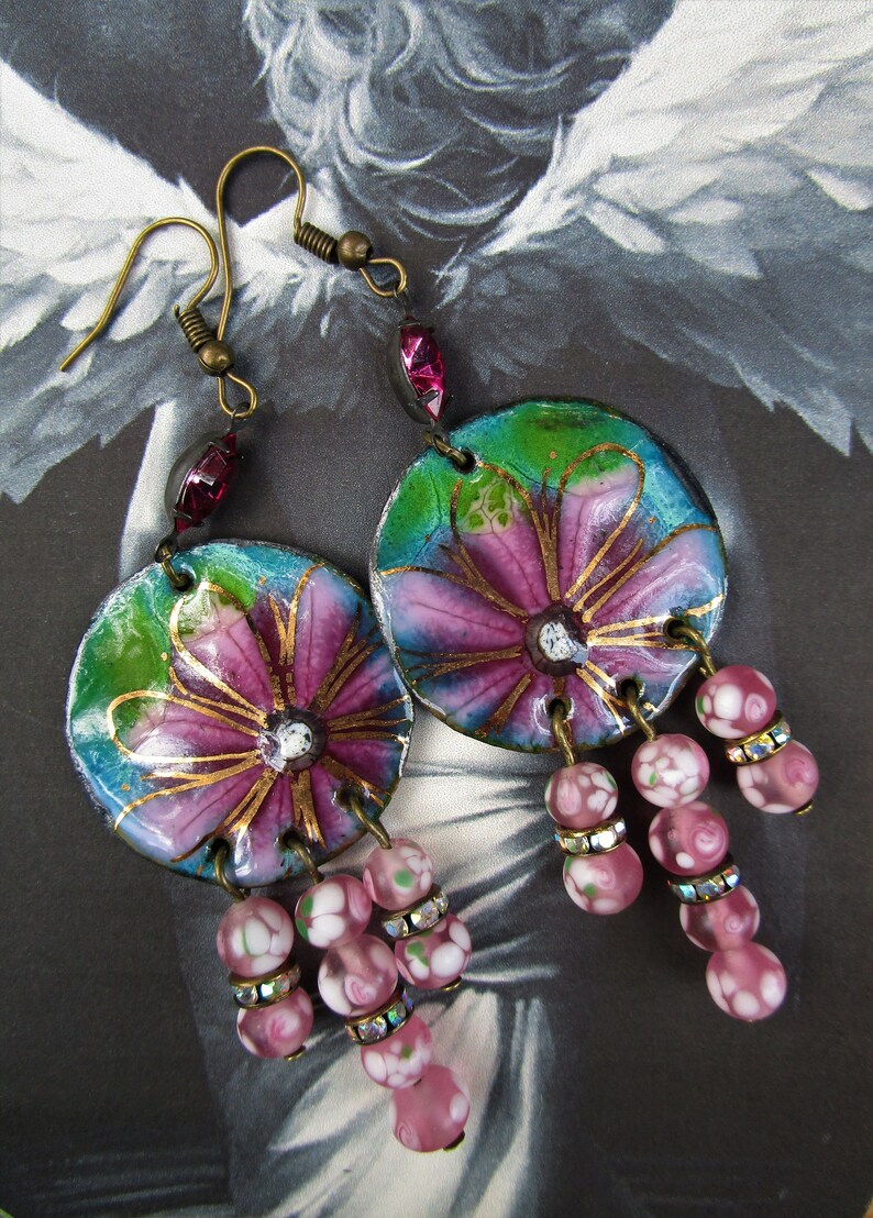 May include: A pair of dangle earrings with pink and green floral designs on the round pendant. The pendants are attached to a chain of pink and white glass beads.