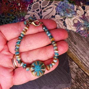 May include: A beaded bracelet with a turquoise and gold-colored central bead. The bracelet features small, round beads in shades of blue, brown, and gold. The clasp is gold-colored. The bracelet is held in a hand.