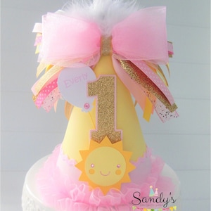 May include: A yellow and pink birthday hat with a gold glitter number one, a sun with a smiling face, and a pink bow with a white feather. The hat says "Everly" on a white tag.