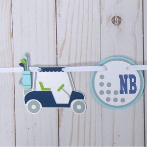 May include: A golf-themed birthday banner with a golf cart, golf balls, and the numbers one and two. The banner is white with blue and green accents. The text on the banner reads 