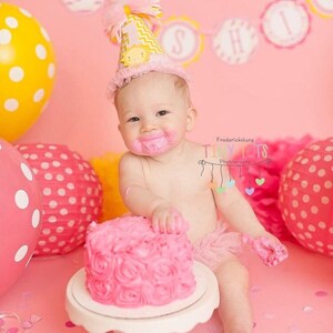 May include: A baby girl wearing a yellow and white chevron birthday hat with a sun and the number one on it, is sitting in front of a pink cake with pink frosting. The baby is wearing a pink tutu and has pink frosting on her face. The background is pink with a yellow and pink banner that says "Sunshine".