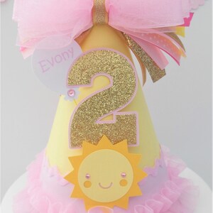 May include: A yellow and pink birthday party hat with a large pink bow, a gold glitter number 2, a yellow sun with a smiling face, and the name "Evony" on a heart-shaped tag. The hat has a pink ruffled trim.
