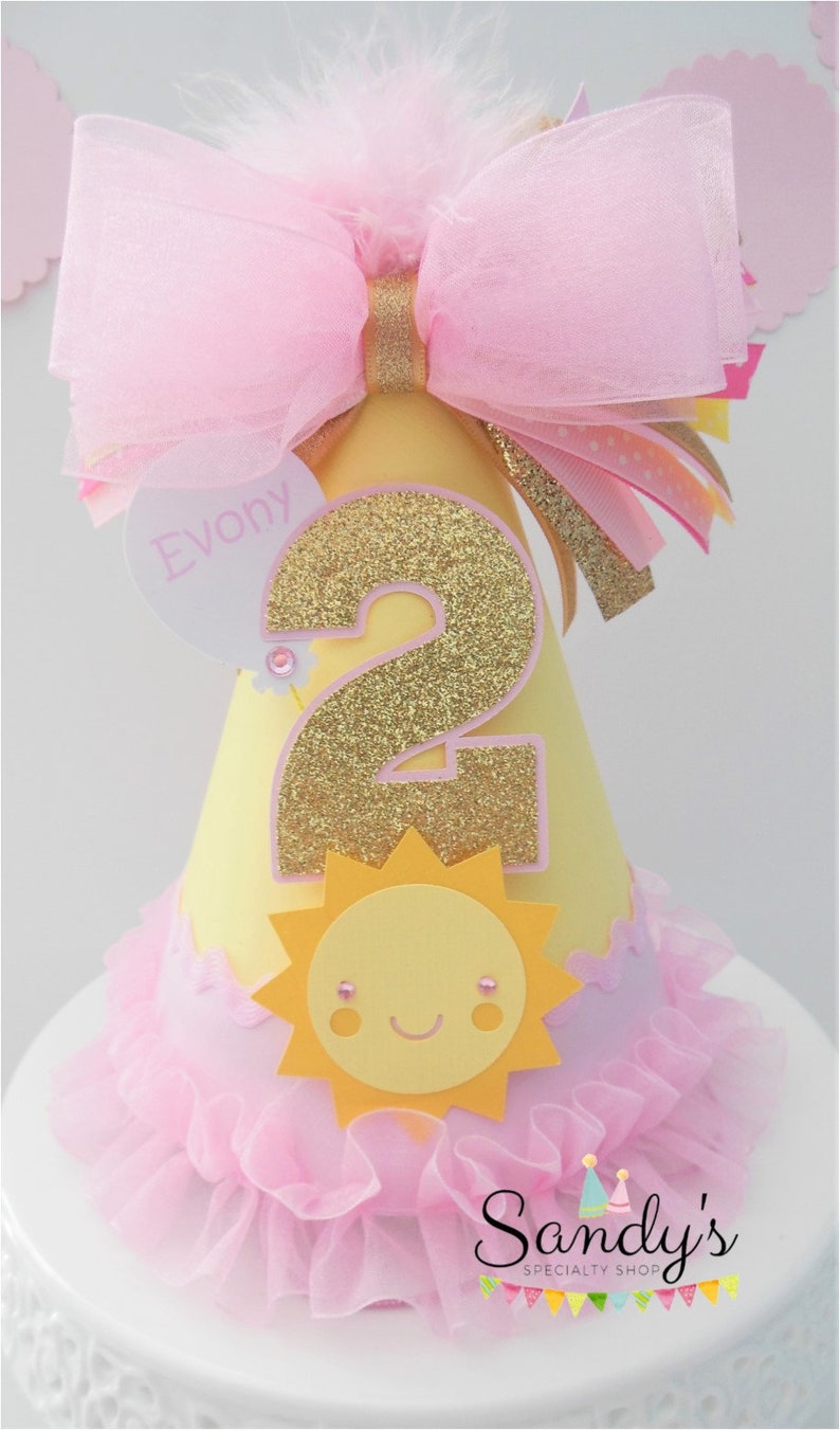 May include: A yellow and pink birthday party hat with a large pink bow, a gold glitter number 2, a yellow sun with a smiling face, and the name "Evony" on a heart-shaped tag. The hat has a pink ruffled trim.