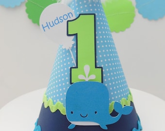 Può includere: Cappello per feste di compleanno blu e verde con pois bianchi, una balena e il numero uno. Il cappello ha anche una piuma bianca in cima e un'etichetta che dice "Hudson".