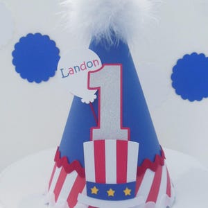 May include: A blue cone-shaped party hat topped with a white feather pom-pom. The hat features a large silver glitter number one, a red and white striped top hat graphic with three yellow stars, and a white balloon cutout with the name Landon.