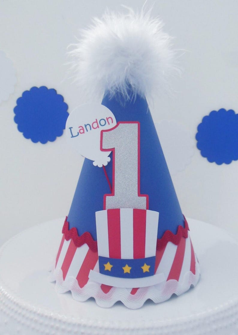 May include: A blue cone-shaped party hat topped with a white feather pom-pom. The hat features a large silver glitter number one, a red and white striped top hat graphic with three yellow stars, and a white balloon cutout with the name Landon.