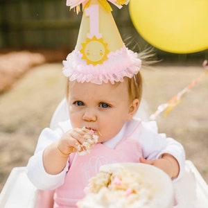May include: A baby wearing a pink and yellow sun-themed birthday hat with the number "1" on it, is eating a pink and white frosted cake.