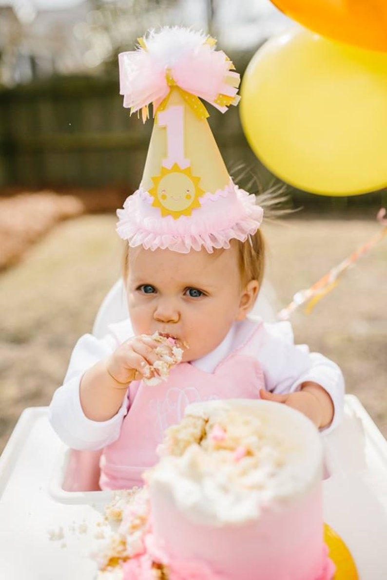 May include: A baby wearing a pink and yellow sun-themed birthday hat with the number "1" on it, is eating a pink and white frosted cake.