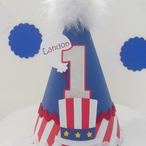 May include: Blue and red birthday party hat with a white feather on top. The hat has a white circle with the name Landon and the number 1 on it. The hat also has a red, white, and blue Uncle Sam hat design.