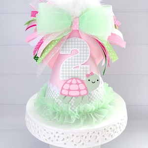 May include: A pink and green birthday party hat with a number 2 and a turtle design. The hat is decorated with a green ruffle, a large bow, and ribbons. It is displayed on a white cake stand.