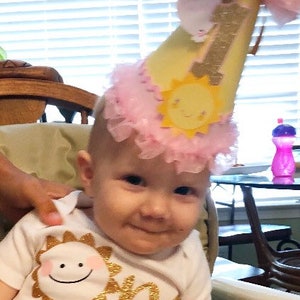 May include: A baby wearing a white onesie with gold glitter lettering that says "One Sunshine" and a yellow and pink birthday hat with a sun design. The baby is holding a piece of food in their hand.