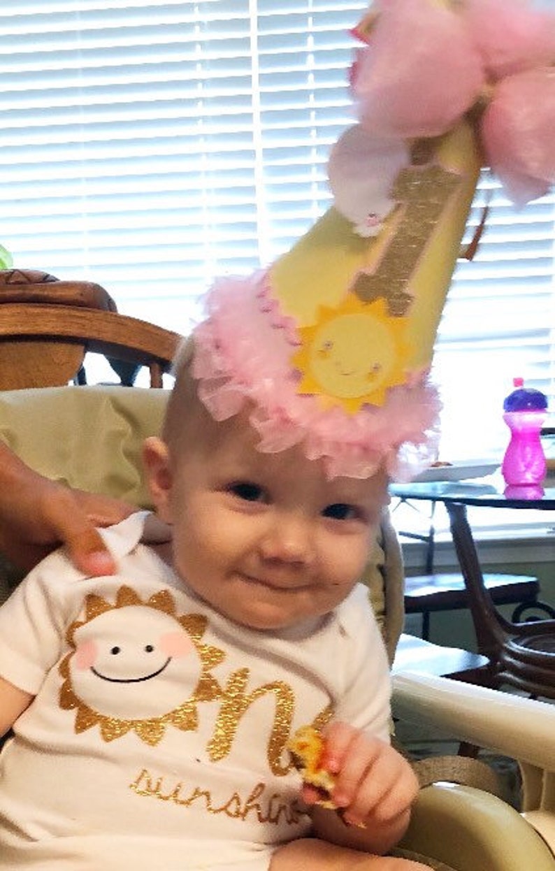 May include: A baby wearing a white onesie with gold glitter lettering that says "One Sunshine" and a yellow and pink birthday hat with a sun design. The baby is holding a piece of food in their hand.