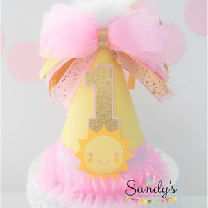 May include: A yellow and pink birthday party hat with a gold glitter number one, a yellow sun with a smiling face, and a pink tulle bow. The hat is on a white cake stand with a pink tulle ruffle.