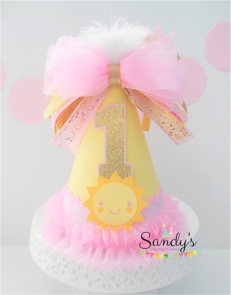 May include: A yellow and pink birthday party hat with a gold glitter number one, a yellow sun with a smiling face, and a pink tulle bow. The hat is on a white cake stand with a pink tulle ruffle.