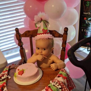 May include: A baby girl wearing a pink and green strawberry-themed birthday hat sits in a high chair with a pink frosted cake in front of her.  The cake is decorated with a strawberry and green leaves.  There are pink and white balloons in the background.