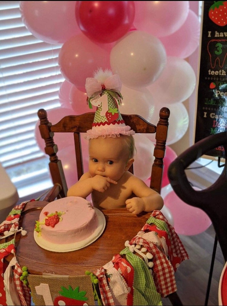 May include: A baby girl wearing a pink and green strawberry-themed birthday hat sits in a high chair with a pink frosted cake in front of her.  The cake is decorated with a strawberry and green leaves.  There are pink and white balloons in the background.