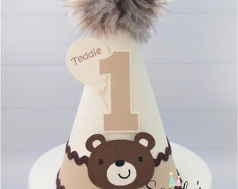 May include: A conical party hat in cream and tan featuring a brown bear face cutout, a large tan number one, and a balloon-shaped tag with the name Teddie printed on it. The top is adorned with a fluffy, multi-toned brown faux fur pom-pom.