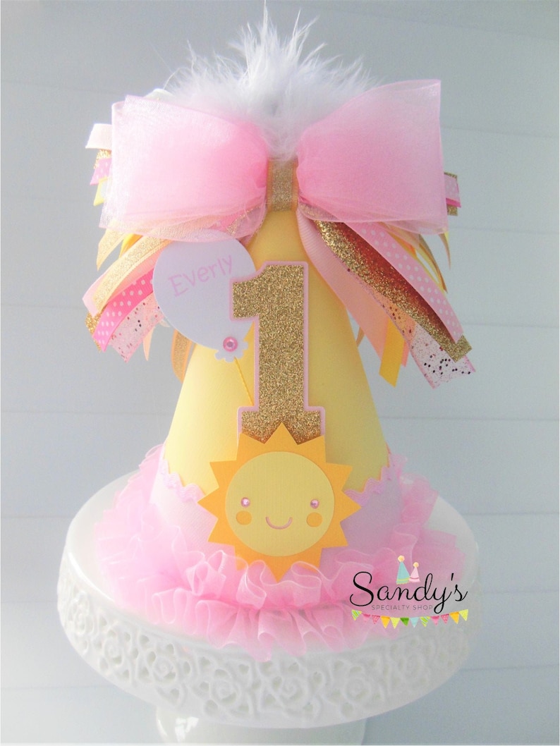 May include: A yellow and pink birthday hat with a gold glitter number one, a sun with a smiling face, and a pink bow with a white feather. The hat says "Everly" on a white tag.