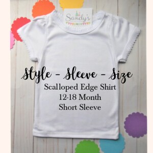 May include: White short-sleeved shirt with scalloped edges and a lace trim around the neckline. The shirt is labeled "Sandy's" and says "Style - Sleeve - Size Scalloped Edge Shirt 12-18 Month Short Sleeve".