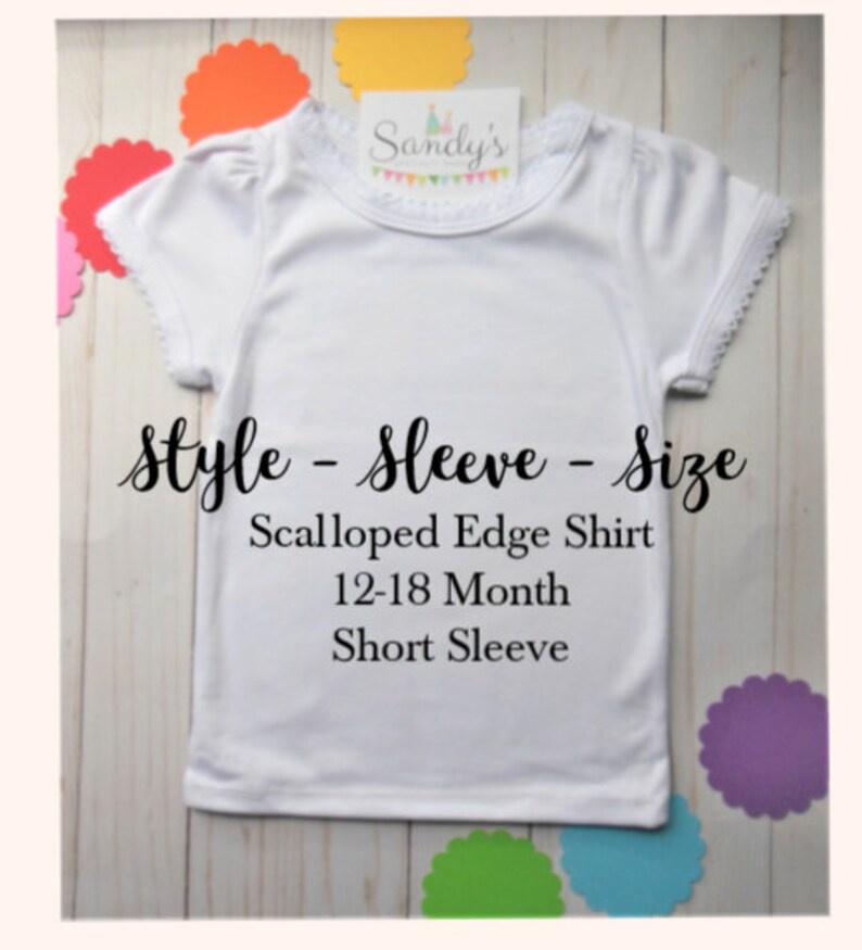 May include: White short-sleeved shirt with scalloped edges and a lace trim around the neckline. The shirt is labeled "Sandy's" and says "Style - Sleeve - Size Scalloped Edge Shirt 12-18 Month Short Sleeve".
