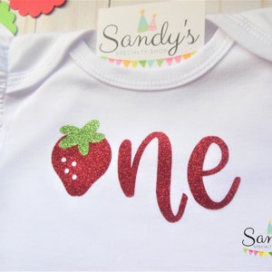 May include: White baby onesie with red glitter 'one' and a red glitter strawberry with green leaves.