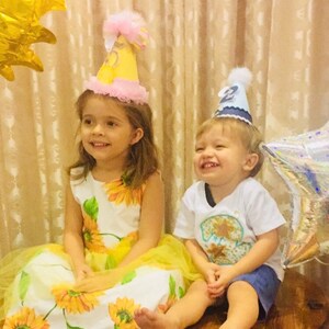 May include: Two children wearing birthday hats sit on a wooden table. The girl is wearing a yellow and white sunflower dress and a pink and yellow hat. The boy is wearing a white shirt with a blue and yellow star design and a blue hat with a white star.