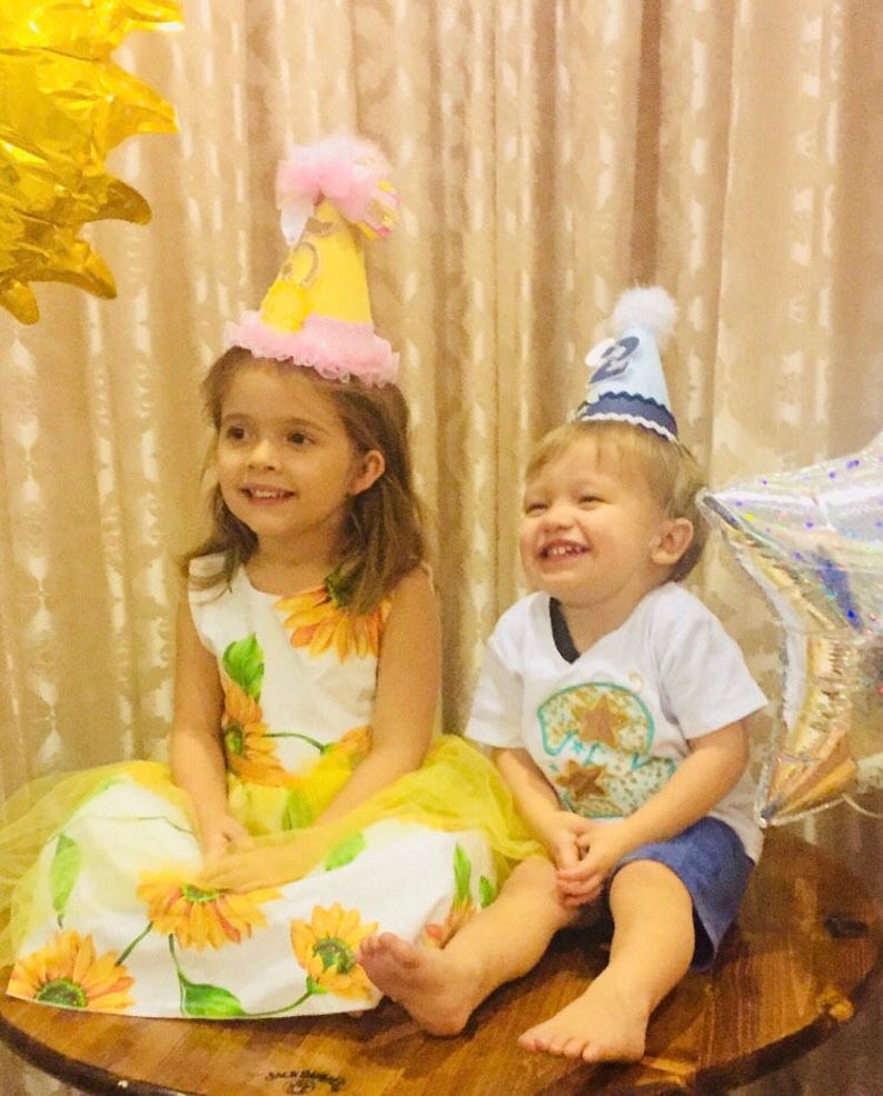 May include: Two children wearing birthday hats sit on a wooden table. The girl is wearing a yellow and white sunflower dress and a pink and yellow hat. The boy is wearing a white shirt with a blue and yellow star design and a blue hat with a white star.