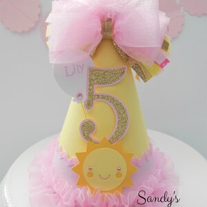May include: Yellow and pink birthday party hat with a gold glitter number 5, a pink bow, and a sun with a smiling face. The hat says "Lily" on the side.