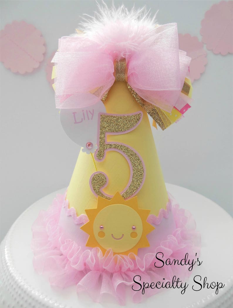 May include: Yellow and pink birthday party hat with a gold glitter number 5, a pink bow, and a sun with a smiling face. The hat says "Lily" on the side.
